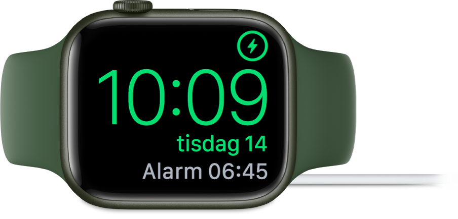 En Apple Watch som ligger på sidan och är ansluten till laddaren. Skärmen visar laddningssymbolen i det övre högra hörnet, under det tiden och sedan tiden för nästa alarm.