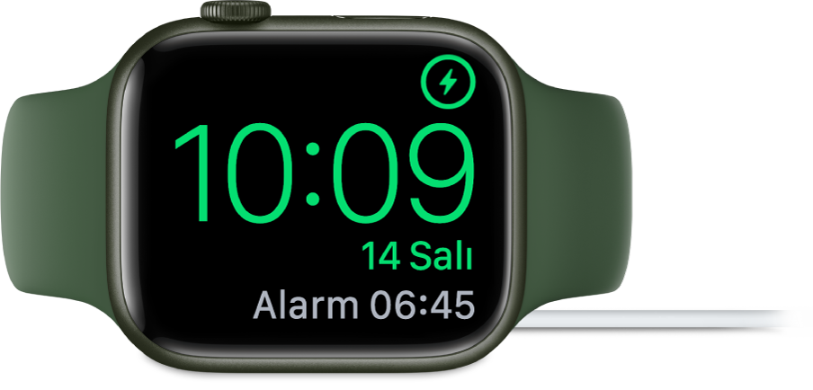 Sağ üst köşede şarj sembolünü, onun altında güncel saati ve bir sonraki alarmın saatini gösteren ekranıyla şarj aletine bağlı olarak yan yerleştirilmiş bir Apple Watch.