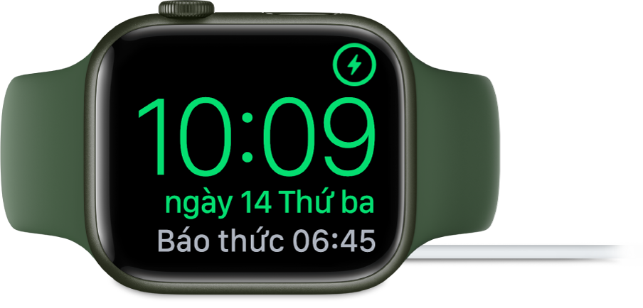 Một Apple Watch được đặt nghiêng và được kết nối với bộ sạc, với màn hình đang hiển thị biểu tượng đang sạc ở góc trên cùng bên phải, thời gian hiện tại ở bên dưới và thời gian của báo thức tiếp theo.