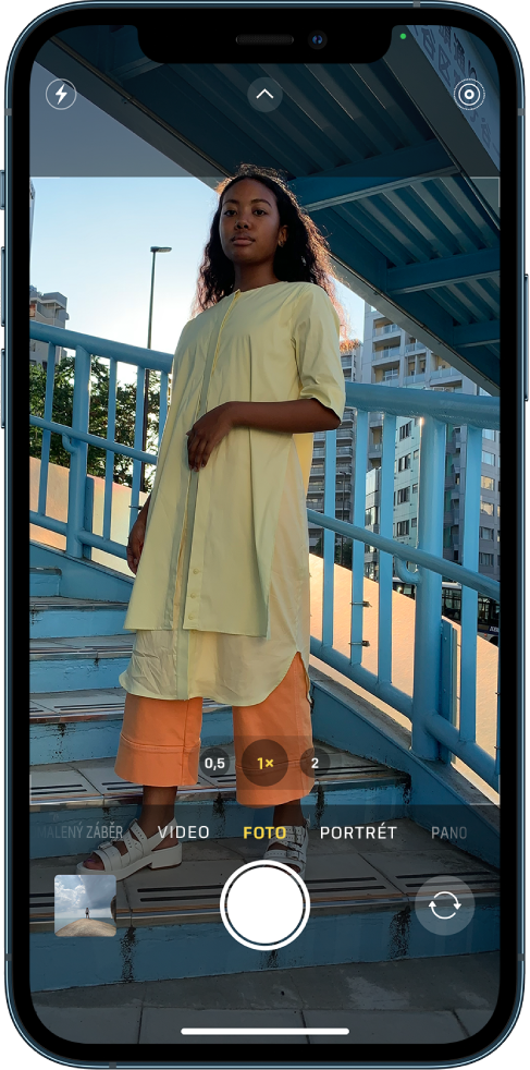 Fotoaparát v režimu Foto s dalšími režimy zobrazenými vlevo a vpravo pod hledáčkem. U horního okraje displeje jsou vidět tlačítka Blesk, Ovládání fotoaparátu a Live Photo. V levém dolním rohu se nachází tlačítko Prohlížeč obrázků a videí. Dole uprostřed je umístěné tlačítko Pořídit snímek a vpravo dole tlačítko přepínače fotoaparátů přepnuté na zadní fotoaparát.
