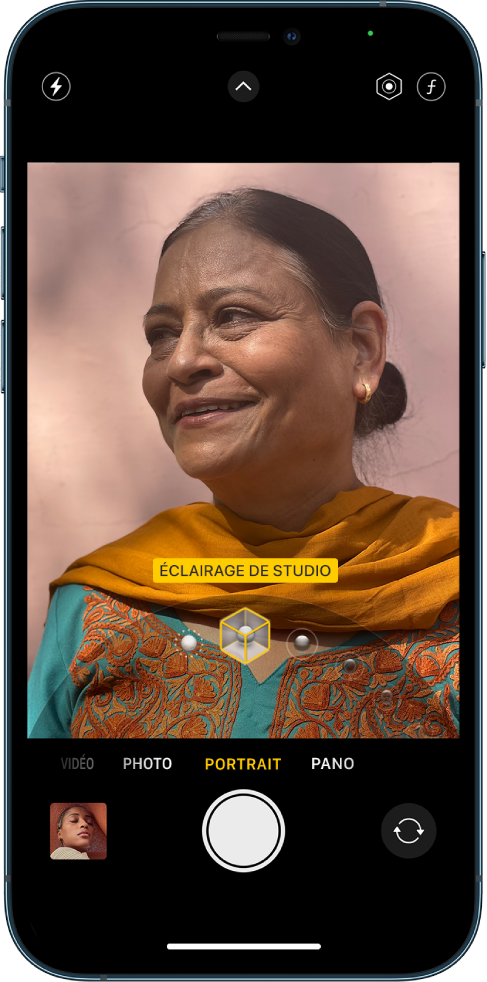 L’écran « Appareil photo » en mode Portrait ; dans le viseur, le sujet est net tandis que l’arrière-plan est flou. Le cadran pour sélectionner les effets d’éclairage Portrait est ouvert en bas de l’image et « Éclairage de studio » est sélectionné. En haut à gauche de l’écran se trouve le bouton Flash, en haut au centre se trouve le bouton « Commandes de l’appareil photo », et en haut à droite de l’écran se trouvent les boutons permettant de régler l’intensité lumineuse du portrait et le contrôle de la profondeur. En bas de l’écran, de gauche à droite, se trouvent le bouton « Visualiseur de photos et vidéos », le bouton « Prendre une photo » et le bouton « Sélecteur de caméra - face arrière ».