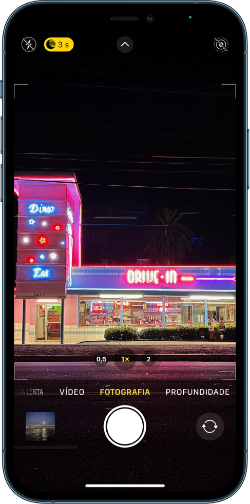 Ecrã da Câmara no modo Fotografia. No canto superior esquerdo do ecrã, estão os botões do flash e do modo Noite. O flash está desativado e o modo Noite está ativado. O botão de controlos da câmara encontra-se na parte superior ao centro e o botão de Live Photo encontra-se no canto superior direito. Na parte inferior do ecrã encontram-se, da esquerda para a direita, o botão de visualização de fotografias e vídeos, o botão para tirar fotografia e o botão de seleção de câmara.