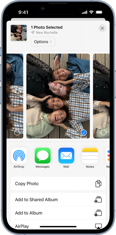 Layar Berbagi dengan foto di sepanjang bagian atas; dua foto dipilih, diindikasikan dengan tanda centang putih dalam lingkaran biru. Baris di bawah foto menampilkan teman yang dapat berbagi dengan Anda menggunakan AirDrop. Di bawahnya terdapat pilihan berbagi lainnya, termasuk, dari kiri ke kanan, Pesan, Mail, Album Bersama, dan Tambahkan ke Catatan. Di baris bawah terdapat tombol Salin, Salin Tautan iCloud, Pertunjukan Slide, AirPlay, dan Tambahkan ke Album.