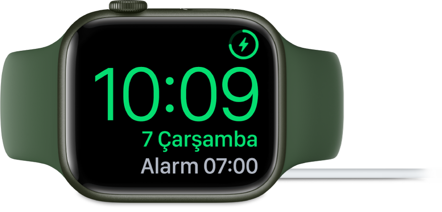 Sağ üst köşede şarj sembolünü, onun altında güncel saati ve bir sonraki alarmın saatini gösteren ekranıyla şarj aletine bağlı olarak yan yerleştirilmiş bir Apple Watch.
