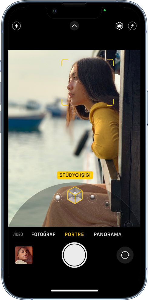 Kamera ekranı Portre modunda; vizördeki konu keskin ve arka plan bulanık. Portre ışıklandırma efektlerinin seçildiği kadran çerçevenin alt kısmında açık ve Stüdyo Işığı seçili. Ekranın sol üst kısmında flaş düğmesi, üst ortada Kamera Denetimleri düğmesi ve ekranın sağ üstünde Portre ışığının yoğunluğunu ve derinlik denetimini ayarlama düğmeleri var. Ekranın en altında, soldan sağa doğru Fotoğraf ve Video Görüntüleyici düğmesi, Resim Çek düğmesi ve Arkaya Bakan Kamerayı Seç düğmesi var.