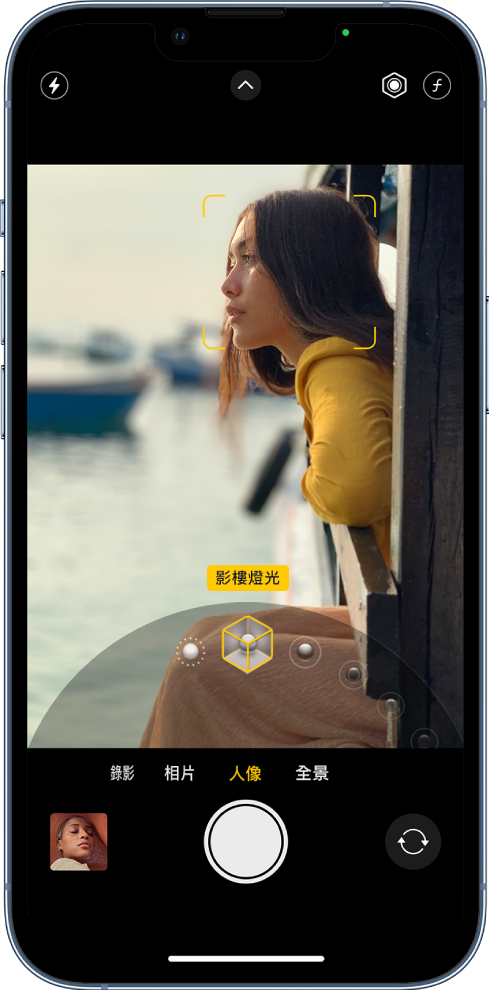 「相機」畫面中的「人像」模式;在觀景器中,主體清晰而背景模糊。用於選擇「人像燈光」效果的刻度盤已在影格下方開啟,並已選擇「影樓燈光」。螢幕左上方是「閃光燈」按鈕,中間上方是「相機控制項目」,而右上方是調整「人像」燈光強度及景深控制的按鈕。螢幕底部由左至右為:「相片和影片檢視器」按鈕、「影相」按鈕和「相機選擇器後置鏡頭」按鈕。