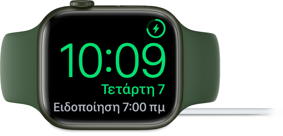 Ένα Apple Watch τοποθετημένο στο πλάι του και συνδεδεμένο στον φορτιστή του. Στην οθόνη εμφανίζεται το σύμβολο φόρτισης στην πάνω δεξιά γωνία, η τρέχουσα ώρα από κάτω η ώρα του επόμενου ξυπνητηριού.