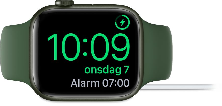 En Apple Watch som ligger på siden og er koblet til laderen, med skjermen som viser ladesymbolet øverst til høyre, gjeldende klokkeslett og tidspunktet for neste alarm.