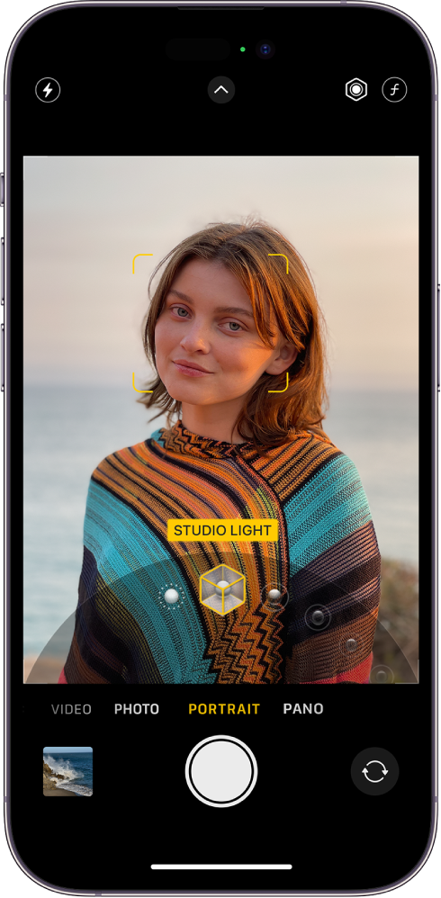 The Camera screen in Portrait mode; in the viewfinder, the subject is sharp and the background is blurred. The dial to select Portrait Lighting is open in the bottom of the frame and Studio Light is selected. At the top left of the screen is the Flash button, at the top center is the Camera Controls button, and at the top right are the buttons to adjust Portrait Lighting intensity and Depth Control. At the bottom of the screen are, from left to right, the Photo and Video Viewer button, the Take Picture button, and the Camera Chooser Back-Facing button.