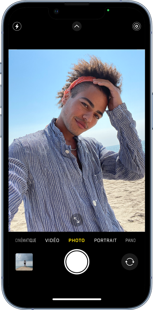 L’écran Appareil photo en mode Photo, avec les autres modes à gauche et à droite sous le viseur. En dessous des différents modes d’appareil photo, de gauche à droite, se trouvent le bouton « Visualiseur de photos et vidéos », le bouton « Prendre une photo » et le bouton « Sélecteur de caméra - face arrière ». Les boutons pour le flash, les commandes de l’appareil photo et le mode Live Photo figurent en haut de l’écran. Dans le cadre de l’appareil photo, le sujet prend un selfie.