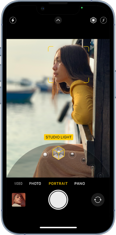 Lietotnes Camera ekrāns režīmā Portrait; skatu meklētājā objekts ir fokusā, un fons ir aizmiglots. Kadra apakšdaļā atvērta skala, kas ļauj izvēlēties režīma Portrait apgaismojuma efektus, un ir atlasīts efekts Studio Light. Ekrāna augšējā kreisajā stūrī ir poga Flash, centrā ir poga Camera Controls, un ekrāna augšējā labajā stūrī ir pogas, kas ļauj pielāgot režīma Portrait apgaismojuma intensitāti un dziļuma vadību. Ekrāna apakšā no kreisās puses uz labo ir pogas Photo and Video Viewer, Take Picture un Camera Chooser Back-Facing.