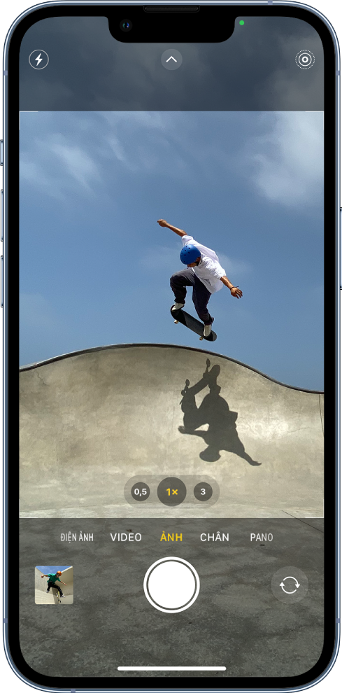 Camera ở chế độ Ảnh, với các chế độ khác ở bên trái và phải bên dưới kính ngắm. Các nút cho Flash, Điều khiển camera và Live Photo xuất hiện ở đầu màn hình. Nút Trình xem ảnh và video ở góc dưới cùng bên trái. Nút Chụp ảnh ở dưới cùng ở giữa và nút Chọn camera mặt sau ở góc dưới cùng bên phải.