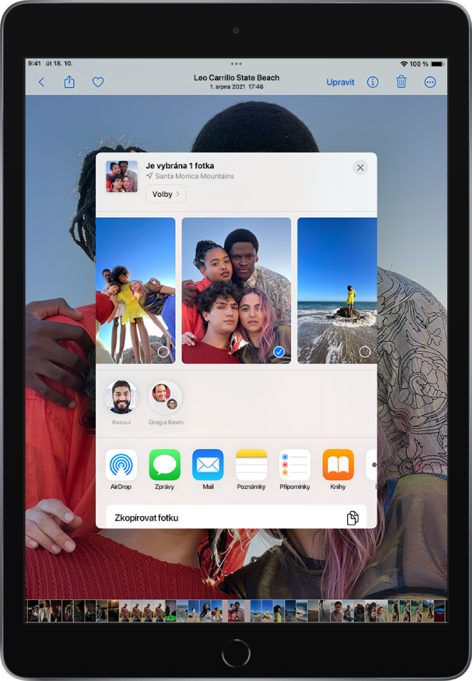 Uprostřed obrazovky se nachází okno sdílení fotek. Podél horního okraje okna jsou rozložené fotky. Jedna z nich je označená zaškrtnutím jako vybraná. Řádek pod fotkami obsahuje návrhy nedávno použitých kontaktů, s kterými můžete fotky sdílet. Pod navrhovanými kontakty se zobrazují volby sdílení, zleva doprava AirDrop, Zprávy, Mail, Poznámky, Připomínky a Knihy. V řádku u dolního okraje obrazovky sdílení jsou uvedené akce. Shora dolů je vidět položky Zkopírovat fotku a Přidat do sdíleného alba.