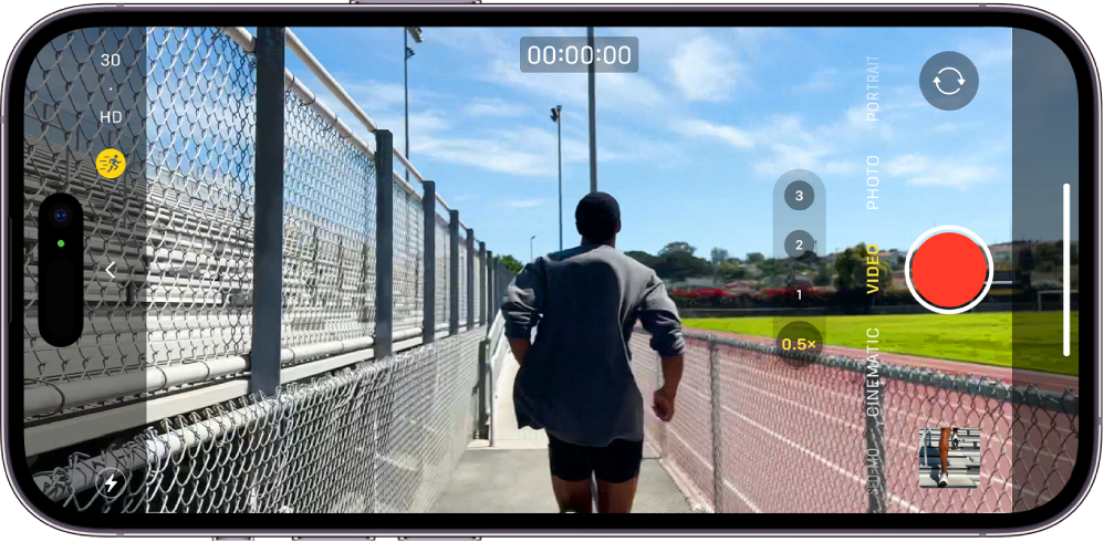 The iPhone is turned horizontally. Camera is open and in Video mode. In the camera frame, a person is jogging. The Flash button is in the bottom-left corner of the screen. The Camera Controls button is in the left-center of the screen. In the top-left corner are quick toggles to switch the video resolution and frame rate. The Action button in the top-left corner is turned on. At the right side are, from top to bottom, the Camera Chooser Back-Facing button, the Record button, and the Photo and Video Viewer button. Within the viewfinder, a person is running.