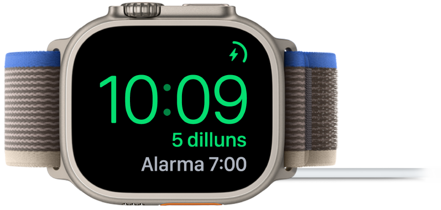 Un Apple Watch posat de costat i connectat al carregador amb la pantalla que mostra el símbol de càrrega a l’angle superior dret, l’hora actual a sota i l’hora de l’alarma següent.