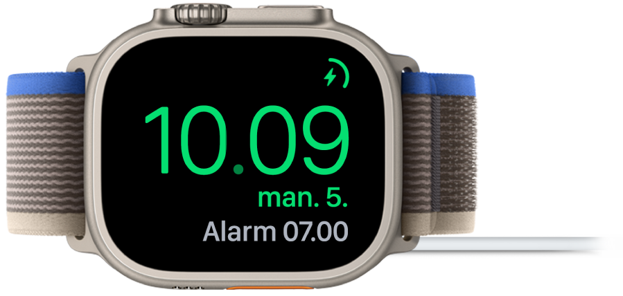 Et Apple Watch, der ligger på siden og er sluttet til opladeren, hvor skærmen viser opladersymbolet øverst til højre, det aktuelle klokkeslæt derunder og klokkeslættet for den næste alarm.