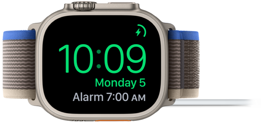 An Apple Watch placed on its side and connected to the charger, with the screen showing the charging symbol in the top-right corner, the current time below that, and the time of the next alarm.