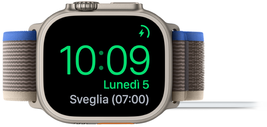 Apple Watch appoggiato su un lato e collegato al caricabatterie, con lo schermo che mostra il simbolo di ricarica nell’angolo superiore destro, l’ora attuale sotto e l’orario della prossima sveglia.