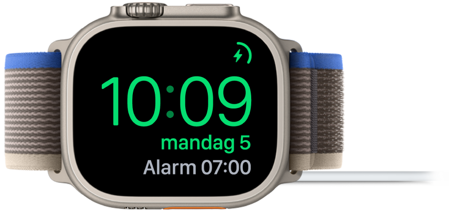 En Apple Watch som ligger på siden og er koblet til laderen, med skjermen som viser ladesymbolet øverst til høyre, gjeldende klokkeslett og tidspunktet for neste alarm.