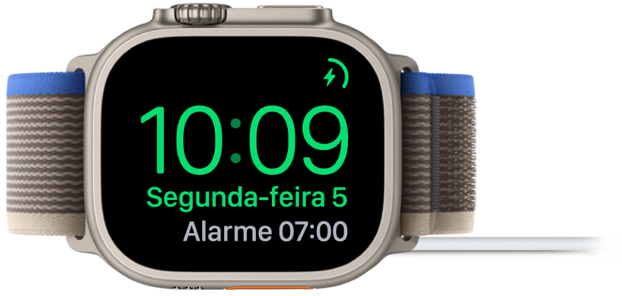 Apple Watch posicionado de lado e conectado ao carregador, com a tela mostrando o símbolo de carregamento no canto superior direito, a hora atual abaixo e o horário do próximo alarme.