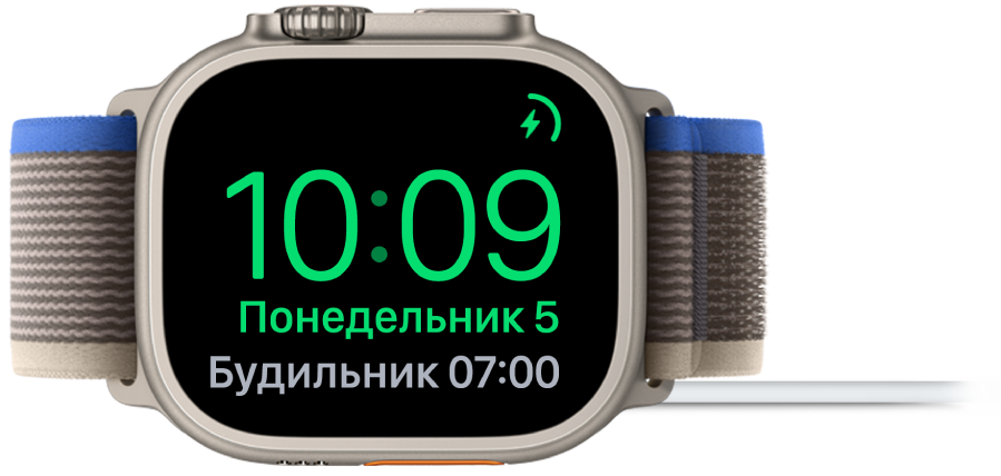 Apple Watch, поставленные набок и подсоединенные к зарядному устройству. На экране отображаются значок зарядки в правом верхнем углу, текущее время под ним и время следующего будильника.