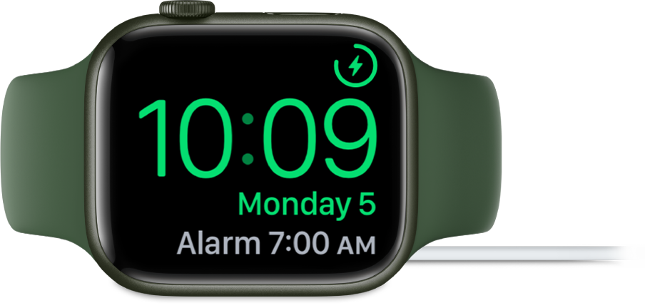 Apple Watch, поставен на една страна и свързан към зарядното устройство, екранът показва символа за зареждане в горния десен ъгъл, текущия час под него и часа на следващата аларма.