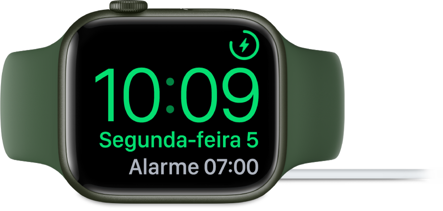 Apple Watch posicionado de lado e conectado ao carregador, com a tela mostrando o símbolo de carregamento no canto superior direito, a hora atual abaixo e o horário do próximo alarme.