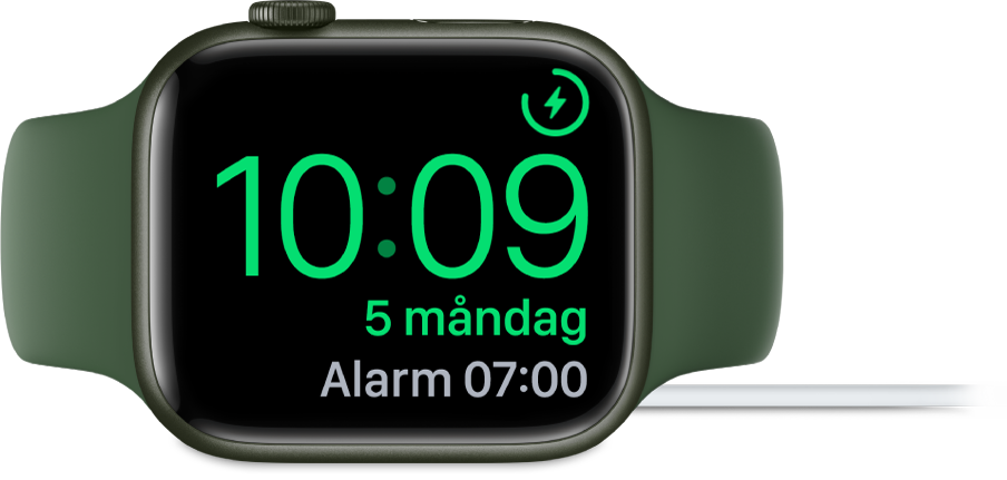 En Apple Watch som ligger på sidan och är ansluten till laddaren. Skärmen visar laddningssymbolen i det övre högra hörnet, under det tiden och sedan tiden för nästa alarm.