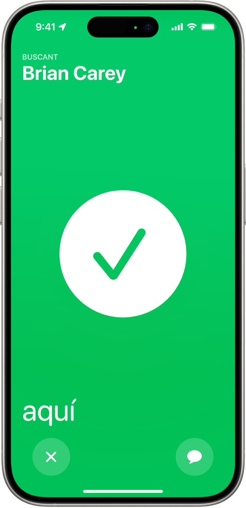 La pantalla de l’iPhone està de color verd i s’hi mostra una marca de verificació al mig. A l’angle superior esquerre hi ha el nom de la persona que s’està buscant i, a l’angle inferior esquerre, la paraula “aquí”, que indica que s’ha trobat.
