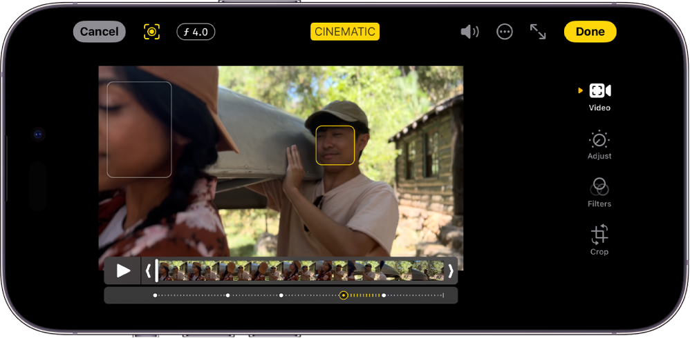Režimo „Cinematic“ redagavimo ekranas gulsčioje padėtyje. Ekrano viršuje yra mygtukas „Cancel“, mygtukas „Cinematic Manual“ ir mygtukas „Depth Adjustment“. Ekrano viršuje centre pasirinktas mygtukas „Cinematic“. Ekrano viršuje dešinėje yra mygtukas „Volume“, mygtukas „More Options“, mygtukas „Enter Full Screen“ ir mygtukas „Done“. Vaizdo įrašas yra ekrano centre, o aplink sufokusuotą objektą yra rėmelis. Po vaizdo įrašu yra kadrų peržiūros priemonė, kuri rodo vaizdo įrašo vietą, kurioje pasikeičia objekto židinys. Redagavimo mygtukai yra dešinėje ekrano pusėje (nuo viršaus į apačią): „Video“, „Adjust Color“, „Filters“ ir „Crop“.