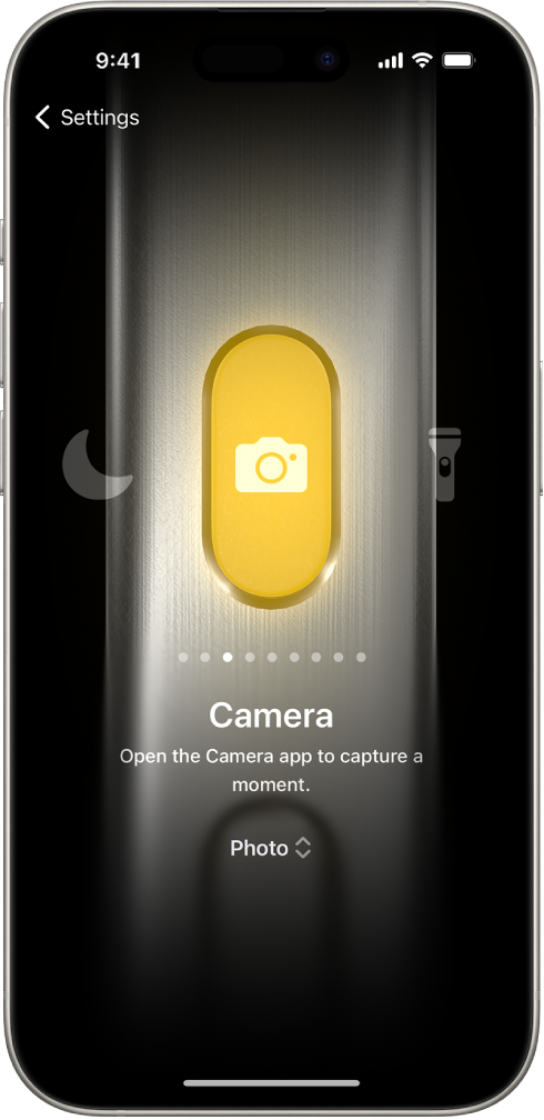 Veiksmo mygtuko individualizavimo ekranas. Pasirinktas veiksmas – fotoaparatas. Kitos funkcijos rodomos dešinėje ir kairėje fotoaparato pusėje, įskaitant „Do Not Disturb“ ir „Flashlight“. Po veiksmais yra taškai, kuriuos palietę galite pereiti prie kito veiksmo. Po pasirinktu veiksmu „Camera“ pateikiamas fotoaparato parinkčių meniu, kurį galite paliesti ir priskirti veiksmo mygtukui.