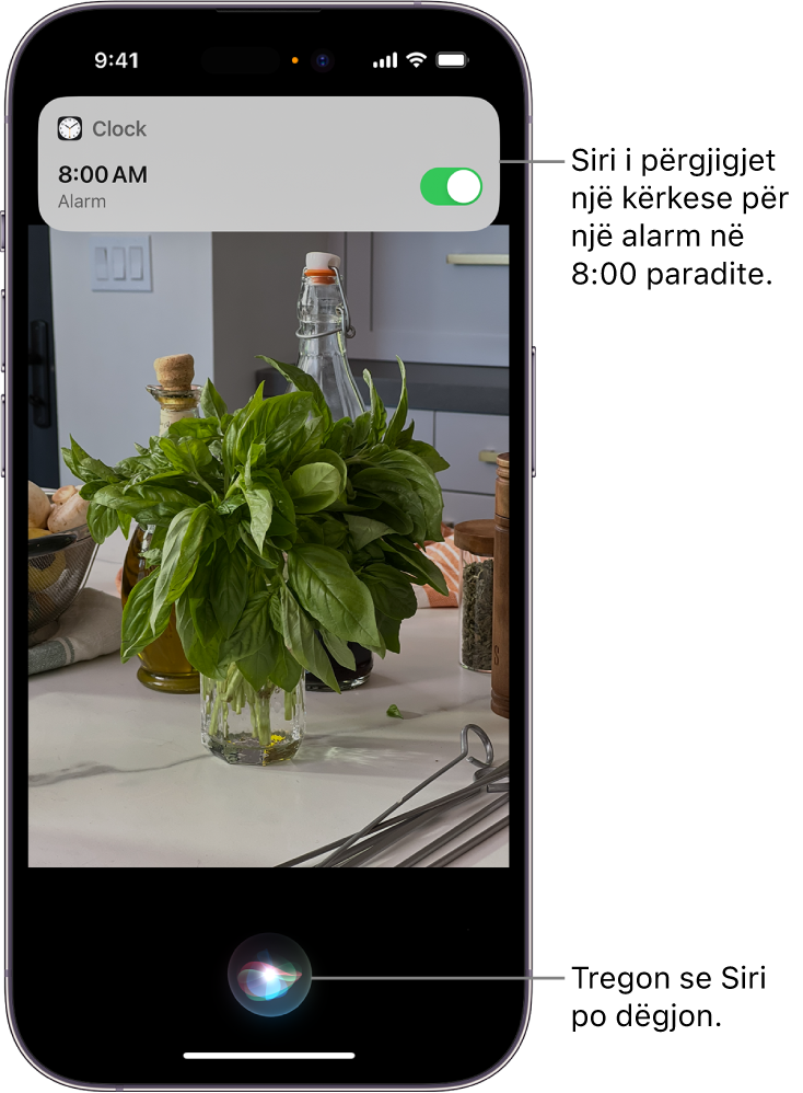Një ekran iPhone. Pranë kreut të ekranit, një njoftim nga aplikacioni Clock tregon se një alarm është i aktivizuar për në orën 8:00 të mëngjesit. Një ikonë në fund të ekranit tregon se Siri është duke dëgjuar.