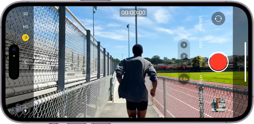 iPhone otočený na šířku. Na displeji je otevřená aplikace Fotoaparát v režimu Video. V levém dolním rohu jsou vidět tlačítka blesku a akčního režimu. Akční režim je zapnutý. Uprostřed levé strany displeje se nachází tlačítko Ovládání fotoaparátu a v levém horním rohu rychlé přepínače, které mění rozlišení videa a snímkovou frekvenci. Po pravé straně je shora dolů rozmístěné tlačítko přepínače fotoaparátů přepnuté na zadní fotoaparát, tlačítko Záznam a tlačítko Prohlížeč obrázků a videí. Fotoaparát zabírá běžící postavu.