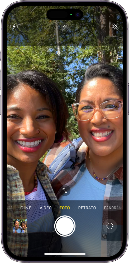La pantalla de la app Cámara en el modo Foto. Debajo de los modos de cámara, de izquierda a derecha, se encuentran los botones Visor de fotos y videos, Tomar foto y Selector de cámara posterior. En la parte superior de la pantalla se encuentran los botones Flash, Controles de la cámara y Live Photos. Dentro del marco de la cámara, las personas se están tomando una selfie.