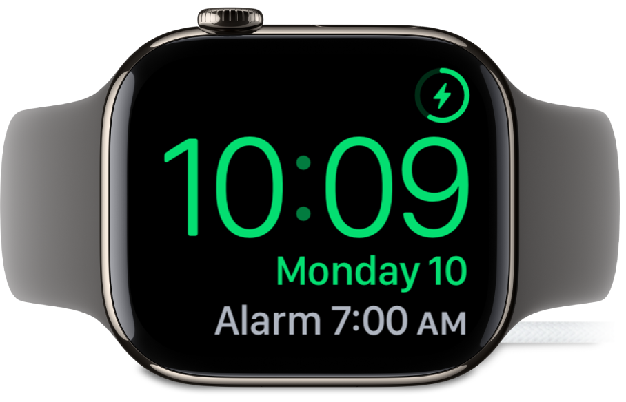 Un Apple Watch colocado de lado y conectado al cargador, con la pantalla mostrando el símbolo de carga en la esquina superior derecha, la hora actual debajo y la hora de la siguiente alarma.