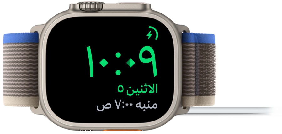 Apple Watch موضوعة على جانبها ومتصلة بالشاحن، وتعرض الشاشة رمز الشحن في الزاوية العلوية اليمنى، والوقت الحالي أسفل ذلك، ووقت المنبه التالي.