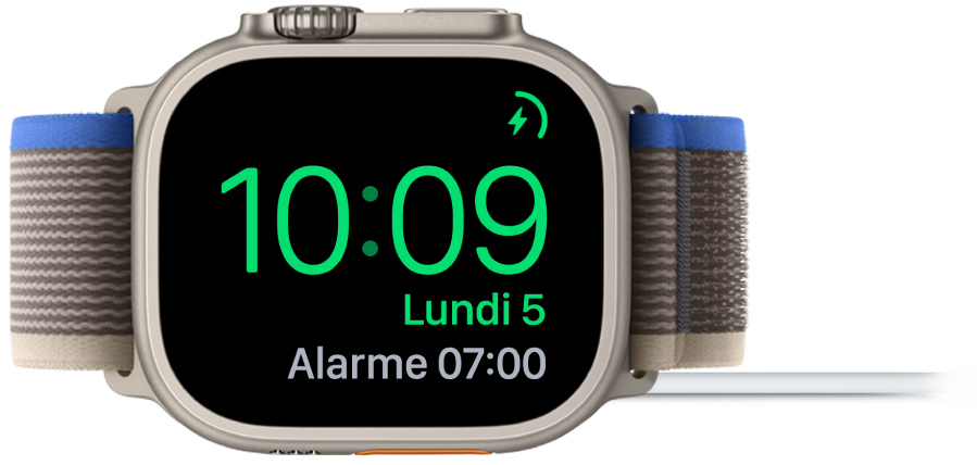 Apple Watch placée sur le côté et connectée au chargeur, dont l’écran affiche le symbole de charge dans le coin supérieur droit, l’heure actuelle en dessous et l’heure de la prochaine alarme.