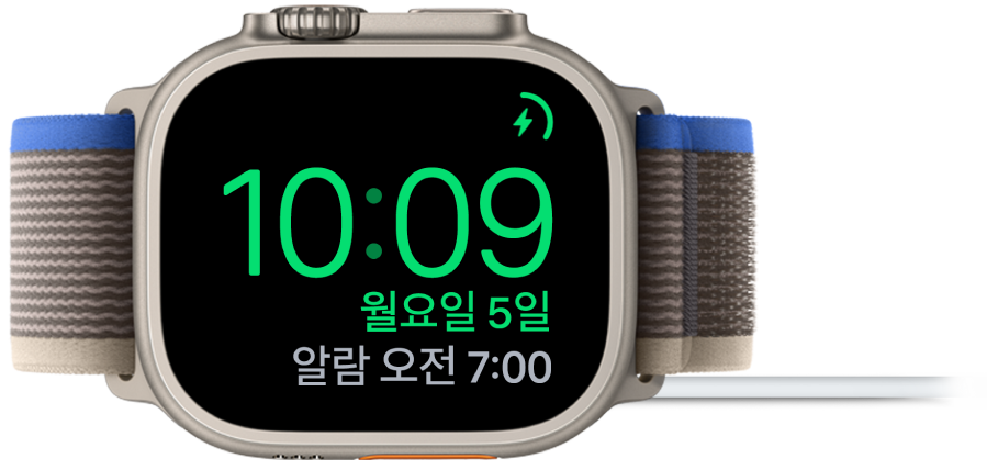 Apple Watch가 옆으로 누워있고 충전기에 연결되어 있음. 화면의 오른쪽 상단 모서리에는 충전 기호가 있고 그 아래에는 현재 시간과 다음 알람 시간이 있음.