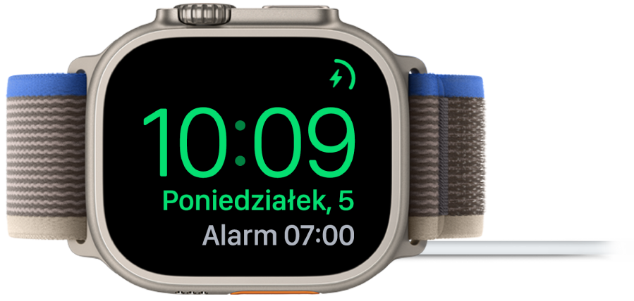 Apple Watch ustawiony na boku i podłączony do ładowarki. W prawym górnym rogu ekranu widoczny jest symbol ładowania, niżej wyświetlana jest bieżąca godzina, a dalej znajduje się czas najbliższego alarmu.