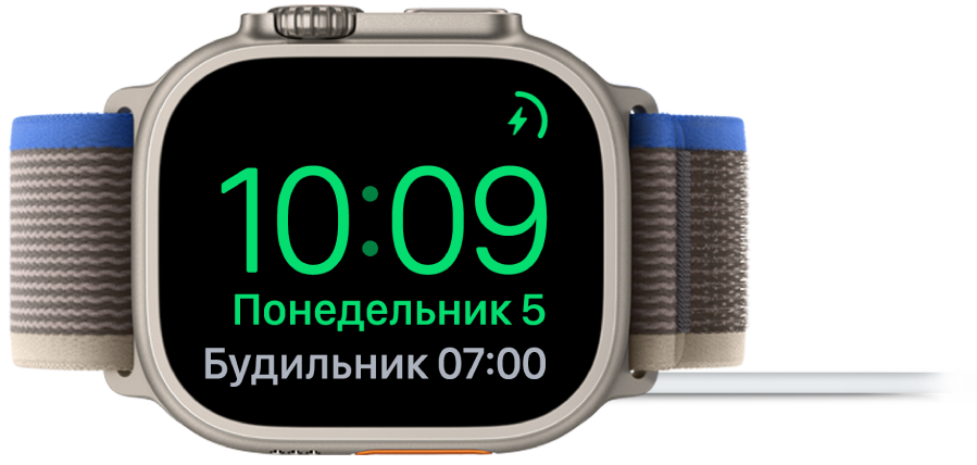 Apple Watch, поставленные набок и подсоединенные к зарядному устройству. На экране отображаются значок зарядки в правом верхнем углу, текущее время под ним и время следующего будильника.