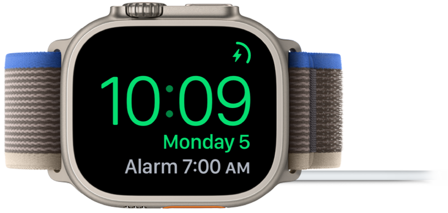 Ura Apple Watch na boku, priklopljena na polnilnik, na zaslonu pa je prikazan simbol za polnjenje v zgornjem levem kotu, trenutni čas pod njim in čas naslednjega alarma.