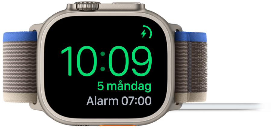 En Apple Watch som ligger på sidan och är ansluten till laddaren. Skärmen visar laddningssymbolen i det övre högra hörnet, under det tiden och sedan tiden för nästa alarm.