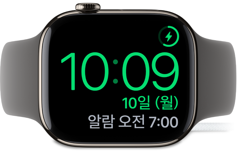 Apple Watch가 옆으로 누워 있고 충전기에 연결되어 있음. 화면의 오른쪽 상단 모서리에는 충전 기호가 있고 그 아래에는 현재 시간과 다음 알람 시간이 있음.