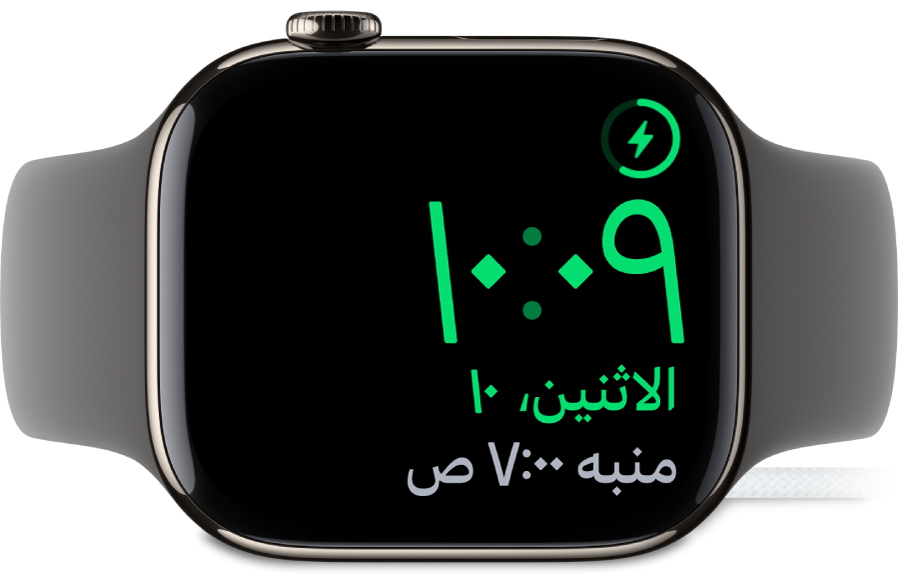 Apple Watch موضوعة على جانبها ومتصلة بالشاحن، وتعرض الشاشة رمز الشحن في الزاوية العلوية اليمنى، والوقت الحالي أسفل ذلك، ووقت المنبه التالي.