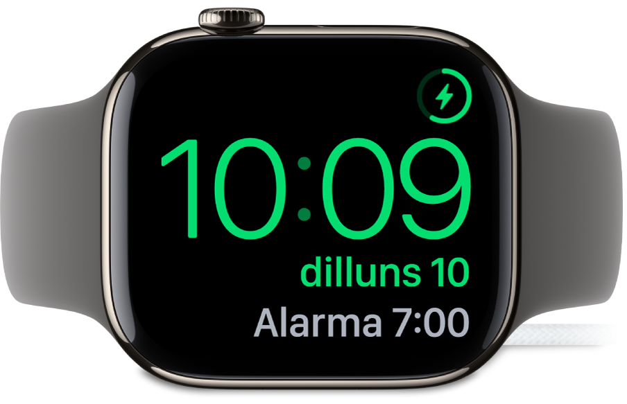 Un Apple Watch posat de costat i connectat al carregador amb la pantalla que mostra el símbol de càrrega a l’angle superior dret, l’hora actual a sota i l’hora de l’alarma següent.