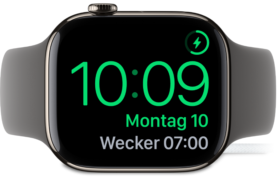 Eine auf die Seite gedrehte Apple Watch, die an das Ladegerät angeschlossen ist und auf deren Display oben rechts das Ladesymbol, darunter die aktuelle Uhrzeit und die Uhrzeit für den nächsten Wecker angezeigt werden.