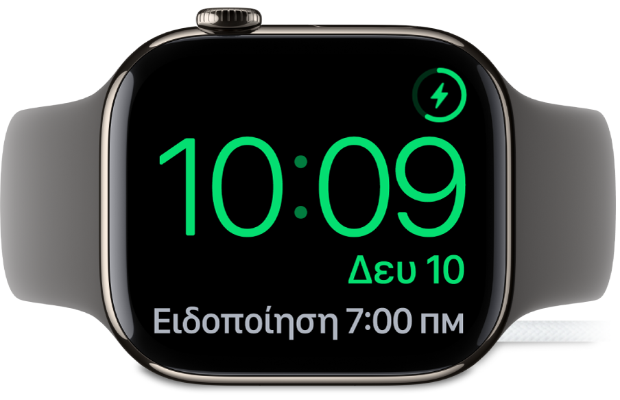 Ένα Apple Watch είναι σε πλάγια θέση και συνδεδεμένο στον φορτιστή του. Στην οθόνη εμφανίζονται το σύμβολο φόρτισης στην πάνω δεξιά γωνία, η τρέχουσα ώρα από κάτω, και η ώρα της επόμενης ειδοποίησης.