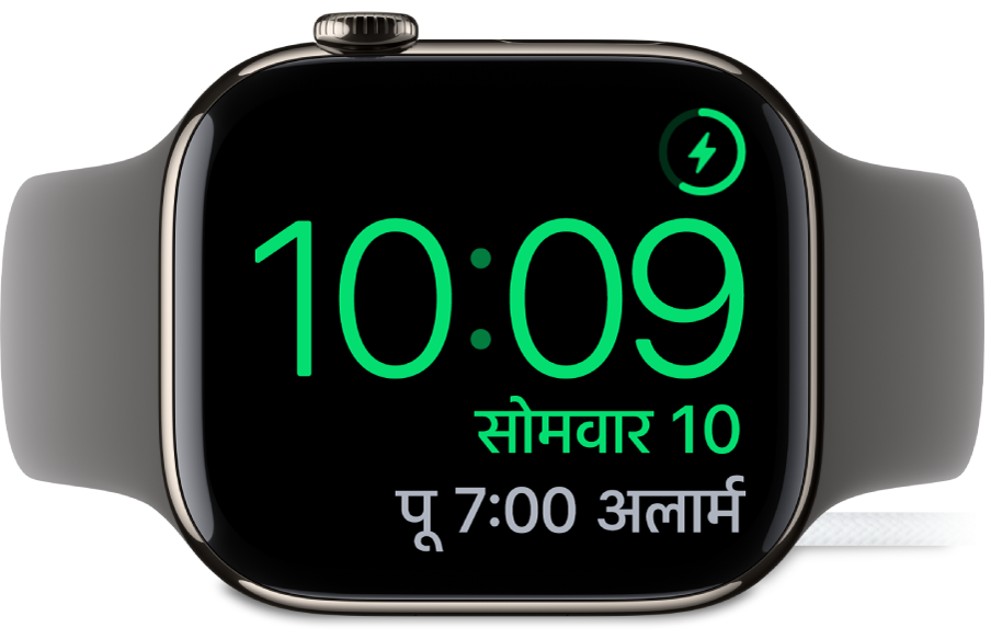 एक Apple Watch जो साइड करके रखी गई है और चार्जर से कनेक्टेड है, जिसमें स्क्रीन के शीर्ष दाएँ कोने में चार्जिंग चिह्न, उसके नीचे वर्तमान समय और अगले अलार्म का समय दिख रहा है।