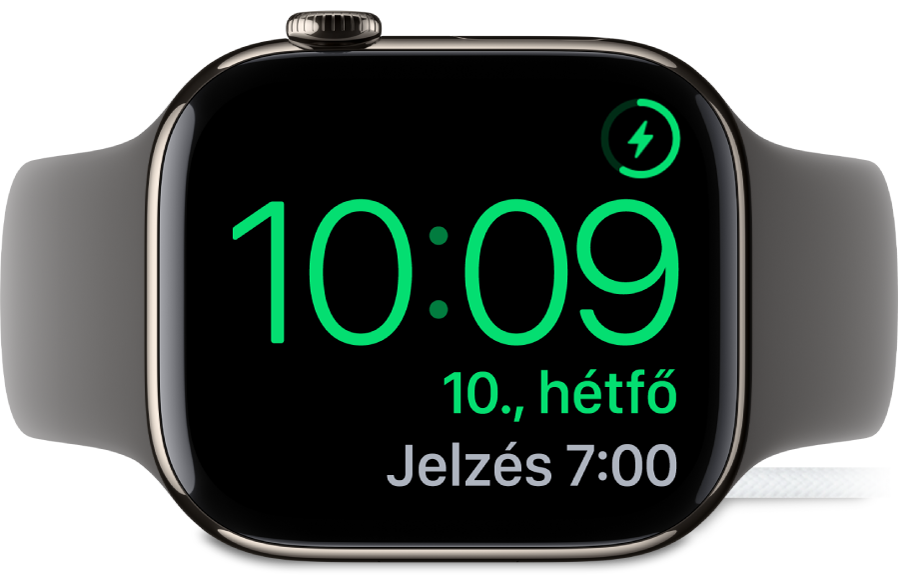 Oldalára fektetett és töltőre csatlakoztatott Apple Watch; a számlap jobb felső sarkában látható a töltés jele, alatta az aktuális idő és a következő jelzés ideje.
