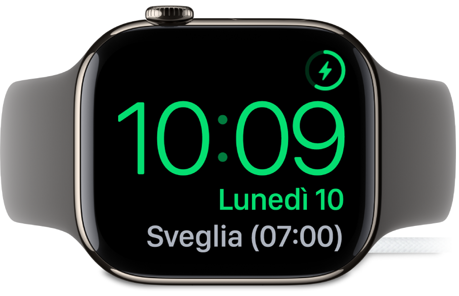 Apple Watch, appoggiato su un lato e collegato al caricabatterie, con lo schermo che mostra il simbolo di ricarica nell’angolo superiore destro, l’ora attuale sotto e l’orario della sveglia successiva.